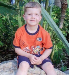 Image of Serese's son, Joseph, a small white boy with blonde hair seated on a rock wearing an orange shirt and blue shorts.